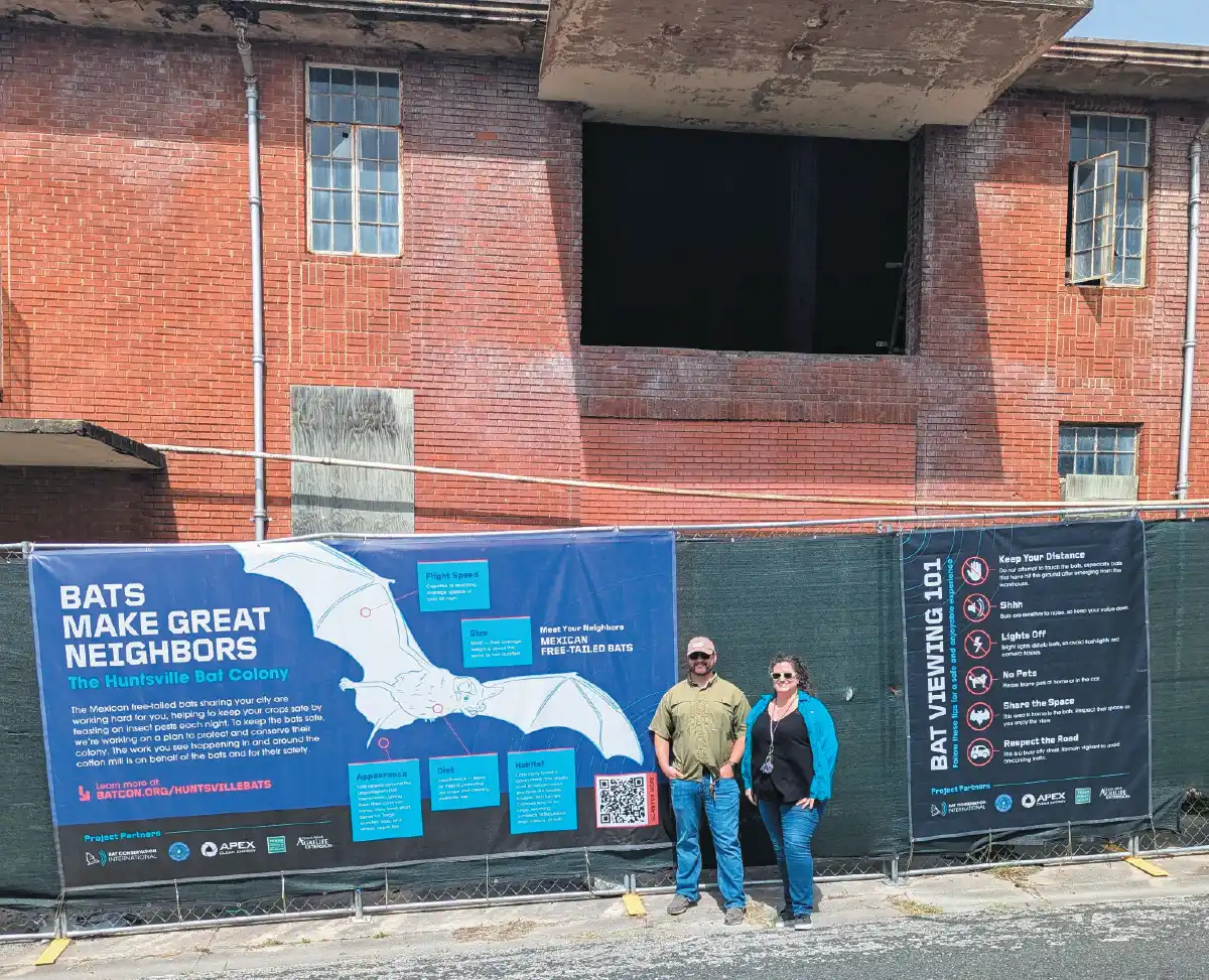Two conservationists standing in front of an educational "Bats Make Great Neighbors" banner at the Huntsville Bat Colony site