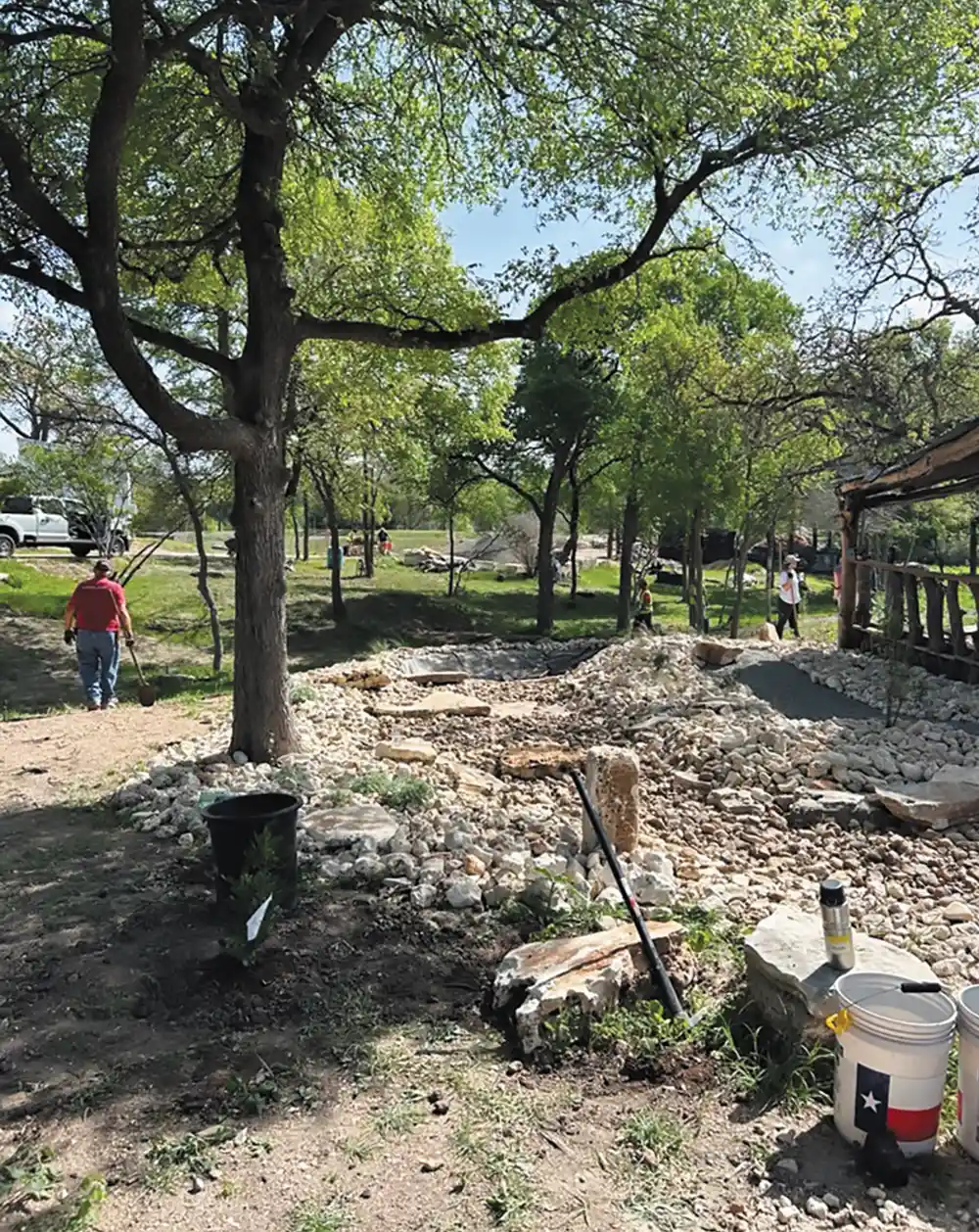 A landscaping crew working on a habitat restoration project, featuring a newly constructed rock garden and drainage area under a canopy of oak trees on a sunny day
