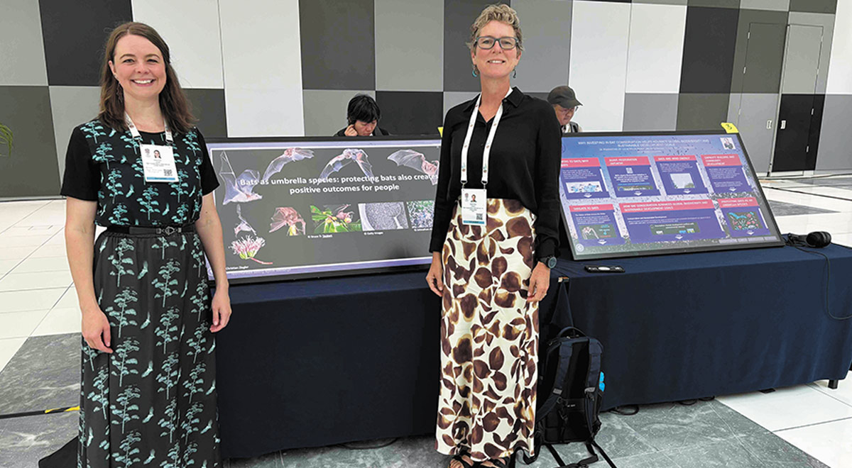 Kristen Lear and Winifred Frick stand behind a table displaying two large posters about bat conservation. One poster reads "Bats as umbrella species." They are in a large hall with a gray and white checkered wall background.