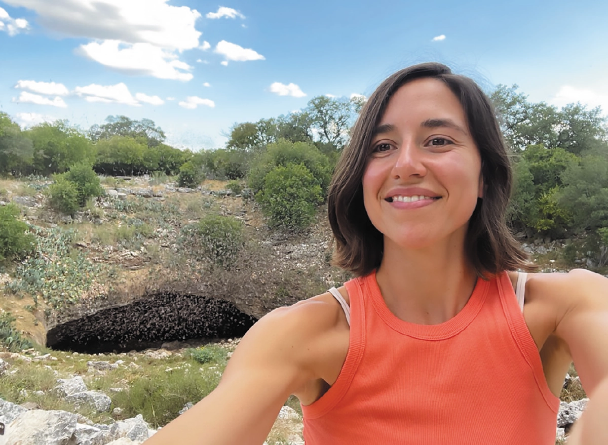 Luz de Wit, Ph.D., a smiling woman in an orange tank top takes a selfie outdoors in a rocky, arid landscape at Bracken Cave in Texas; In the background, a large, dark swarm of bats is visible emerging from or entering a cave opening nestled among green bushes and cacti under a bright, partly cloudy sky