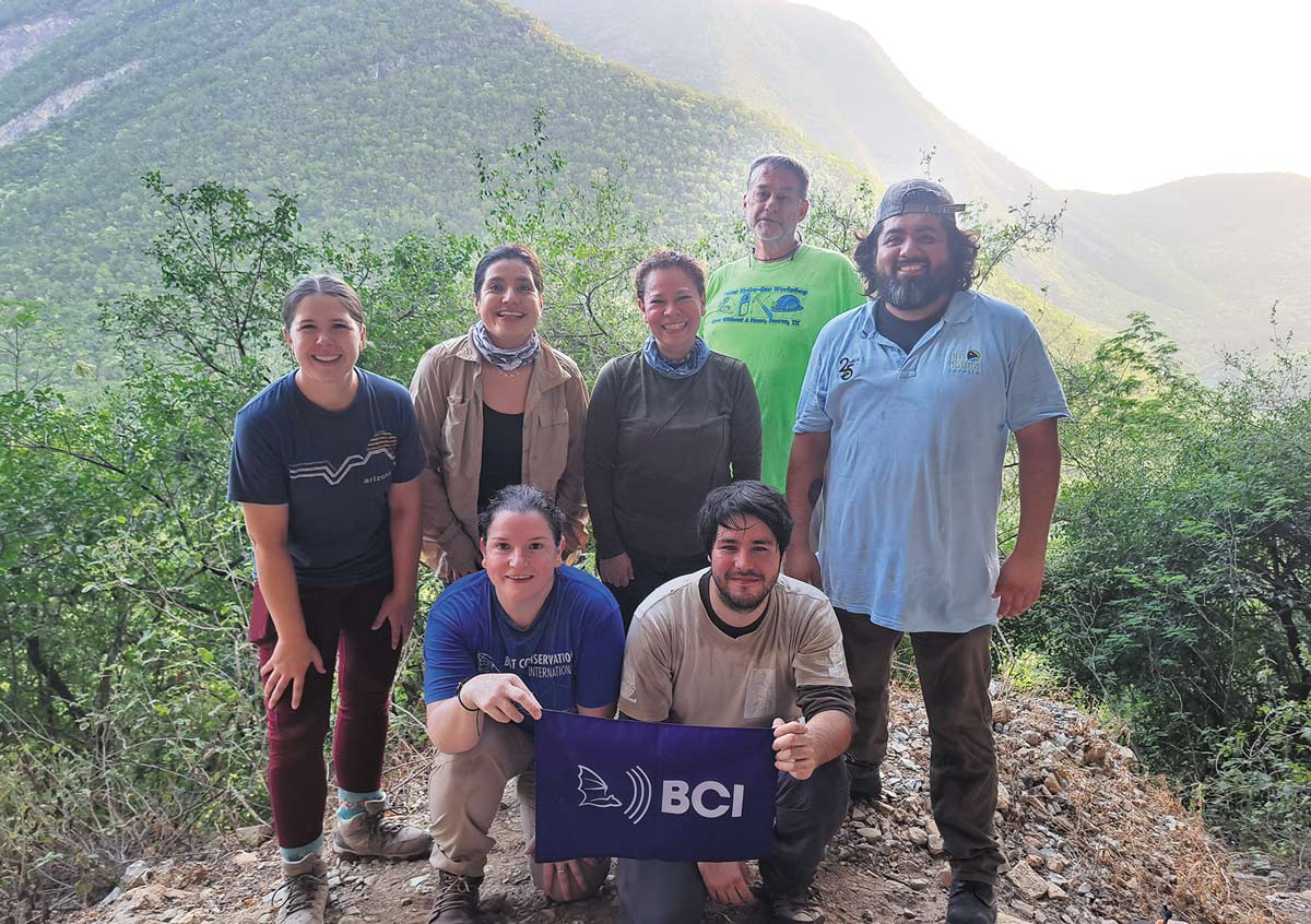 A group of seven researchers poses outdoors on a mountain trail, with two holding a blue Bat Conservation International flag.