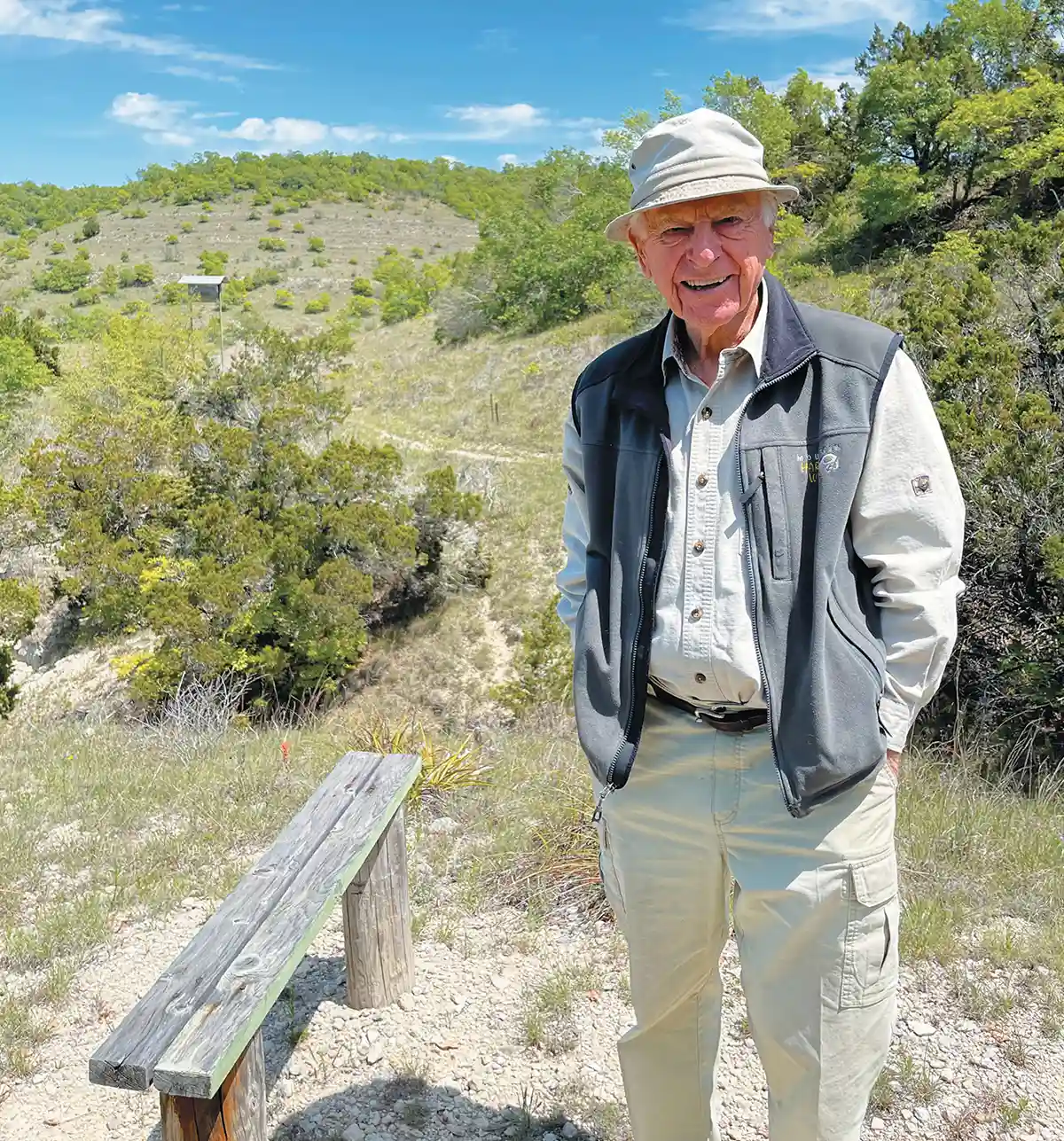 A smiling elderly man wearing a bucket hat and grey vest stands next to a rustic wooden bench on a sunny hillside trail.