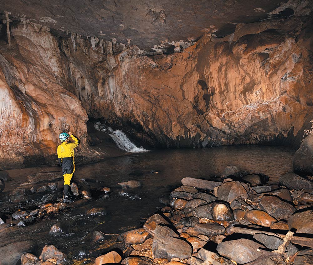 Interior of a limestone cave featuring a subterranean river and a small waterfall and a single explorer looking at the surroundings