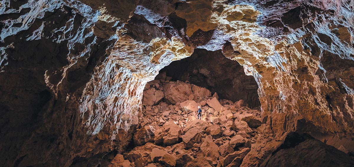 A caver standing among large boulders inside a massive, illuminated underground chamber.