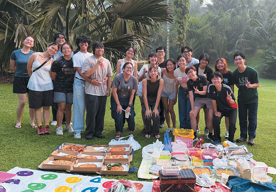 A group of people standing together outdoors behind a picnic spread on the lawn.