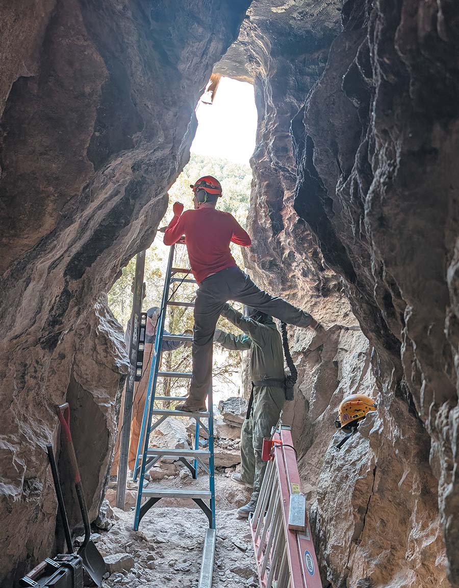 Workers in helmets and protective gear using ladders inside a narrow cave opening.